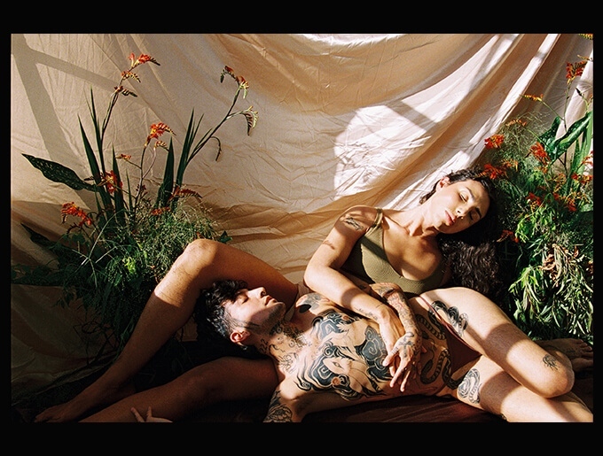 Coyote Park lies on their side, their head facing upward, their head resting between River’s legs. River, sits half-reclined in the opposite direction, their hand entwined with Coyote’s on Coyote’s tattooed chest. Both their eyes are closed. A draped piece of beige fabric fills the background, and the left and right sides of the image have botanical arrangements of greenery with red-orange flowers.